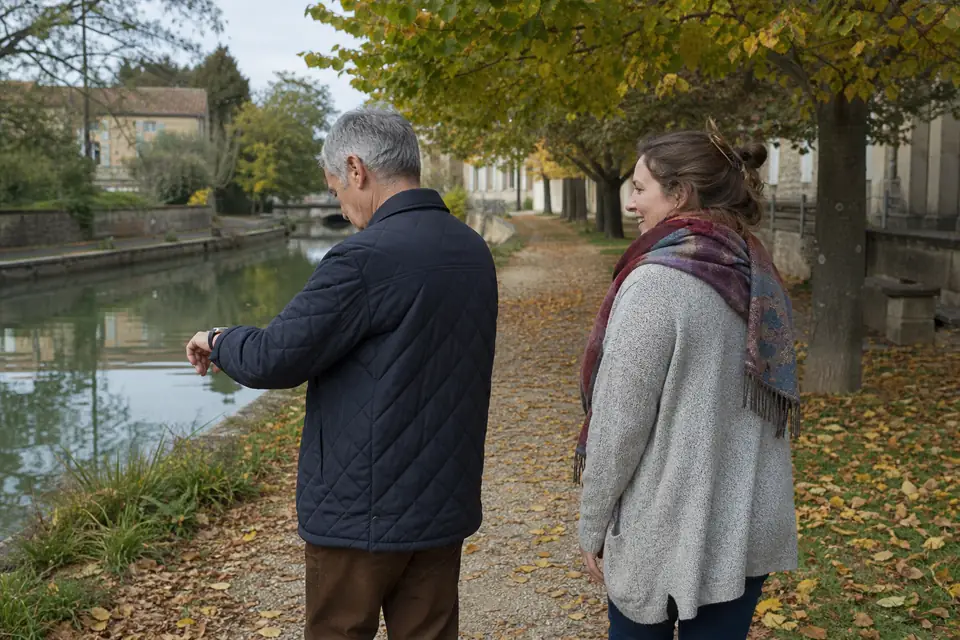 Un homme d’une soixantaine d’années regarde sa montre en marchant avec sa fille sur un chemin de halage bordé d’arbres, un après-midi d’automne.