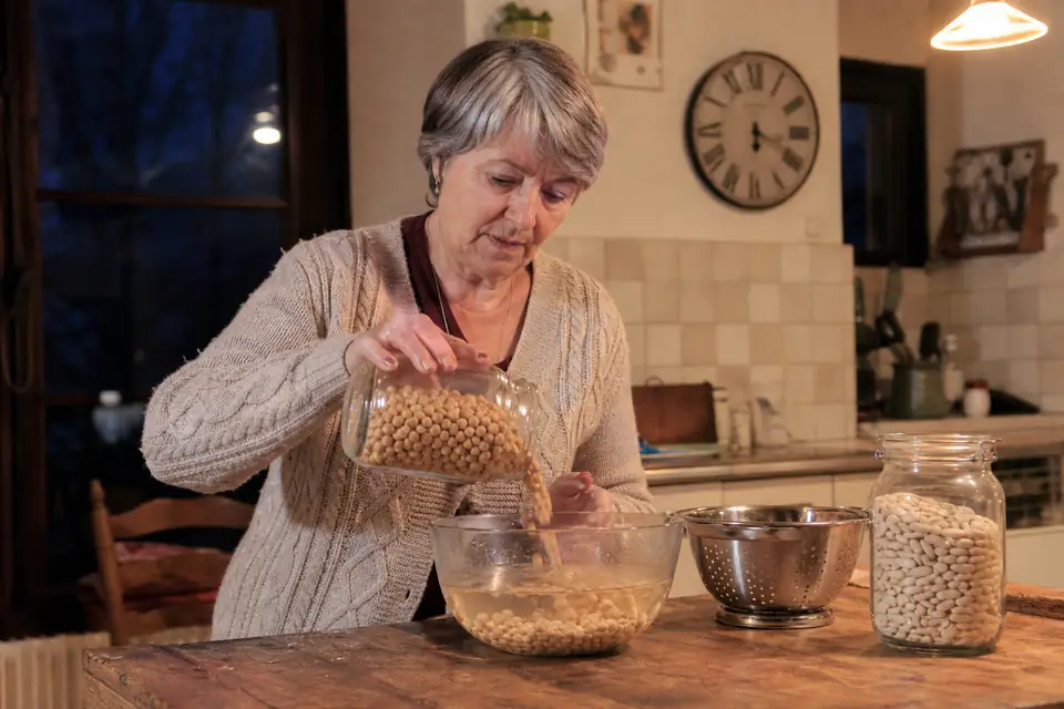 Une femme verse des pois chiches secs dans un grand saladier d’eau froide, à côté d’un bocal de haricots blancs, sur une table de cuisine.